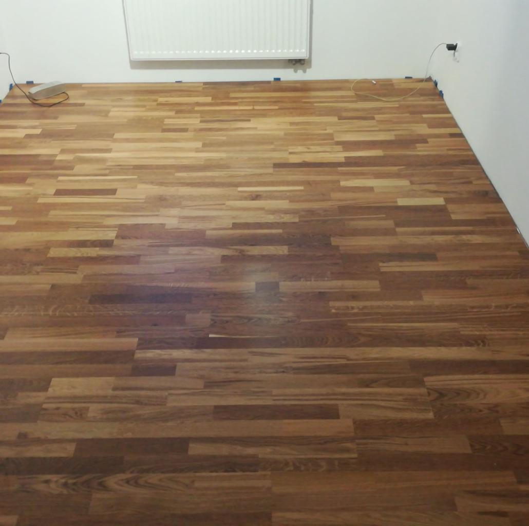 Ułożona, brązowa podłoga z paneli drewnopodobnych w pustym pokoju, zabezpieczona niebieską taśmą malarską przy ścianach, widoczny grzejnik i gniazdko elektryczne.