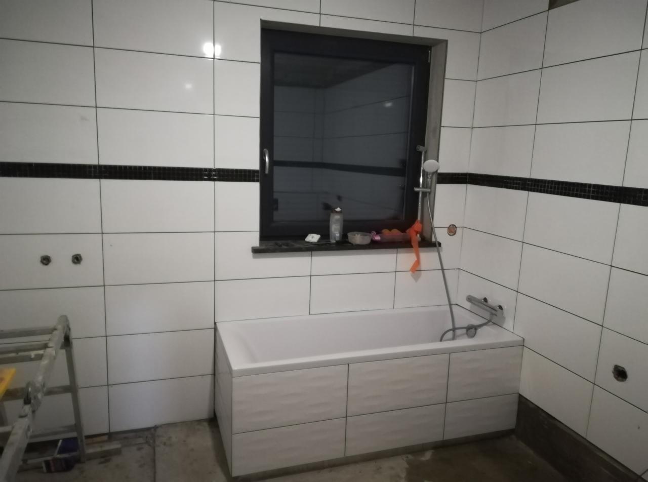 Białe, prostokątne płytki ścienne z czarnym pasem dekoracyjnym, okno z czarną ramą nad wanną z panelami o falistym wzorze, widoczny fragment drabiny malarskiej.