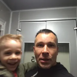 Mężczyzna z synem na tle odnowionego, jasnoszarego pomieszczenia z nowymi drzwiami, sugerującego świeżo zakończony remont. Uśmiechnięta twarz dziecka dodaje pozytywnego akcentu.