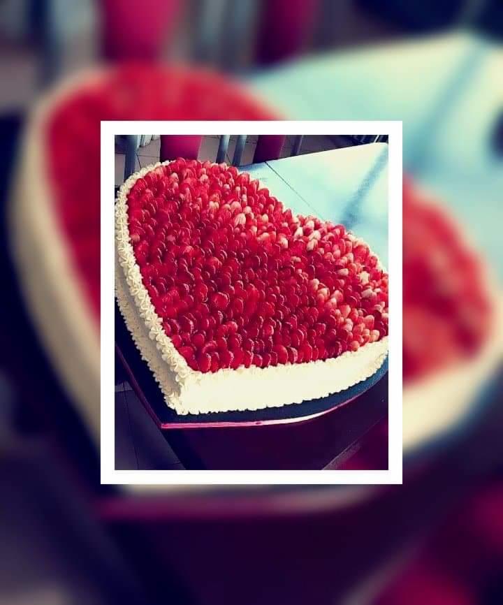 Tort w kształcie serca udekorowany świeżymi truskawkami i kremem, leżący na ciemnym blacie.