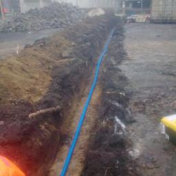 Fosiill - Błękitna rura w wykopie ziemnym, prawdopodobnie instalacja podziemna. W tle budynek, gruz i kontener. Widok na plac budowy w pochmurny dzień.