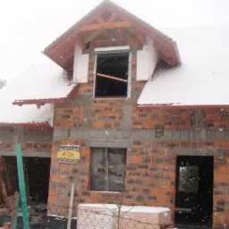 Budowa domu jednorodzinnego w trakcie zimy, widoczne ściany z czerwonej cegły, częściowo ośnieżony dach i okna zabezpieczone folią. Budynek w stanie surowym otwartym.