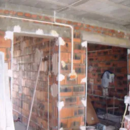 Surowy mur z cegły z widoczną instalacją elektryczną i przygotowanymi otworami na drzwi, w trakcie prac wykończeniowych, z widocznym sprzętem budowlanym.