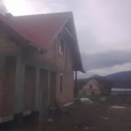 Surowy stan budynku z cegły z czerwonym dachem w pochmurny dzień. Widoczne elementy konstrukcyjne i zarys gór w tle. Etap budowy, widok z zewnątrz.