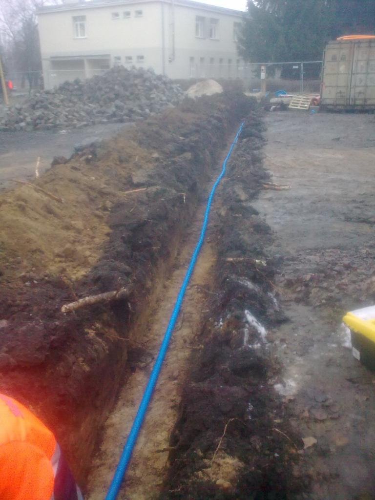 Błękitna rura w wykopie ziemnym, prawdopodobnie instalacja podziemna. W tle budynek, gruz i kontener. Widok na plac budowy w pochmurny dzień.