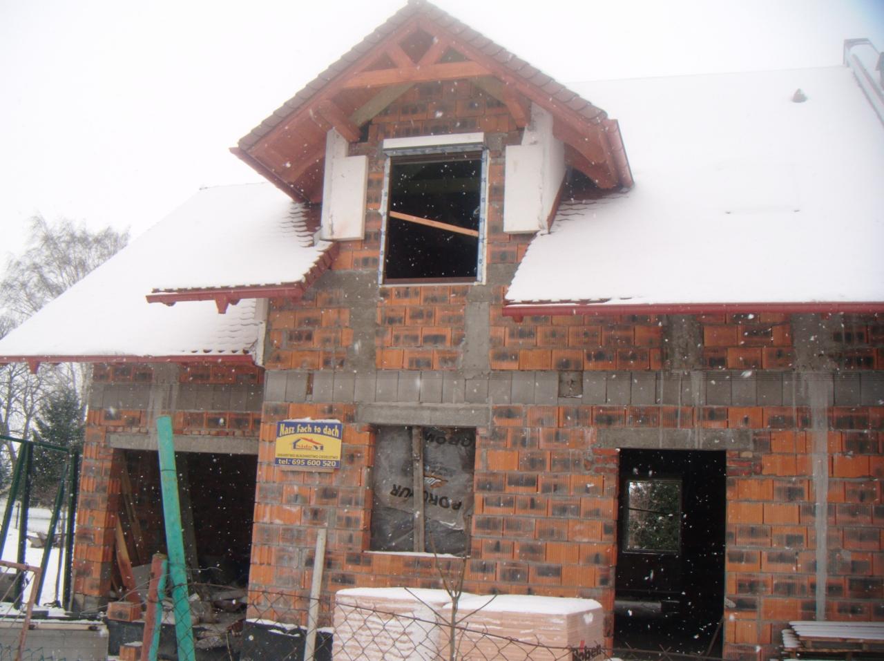 Budowa domu jednorodzinnego w trakcie zimy, widoczne ściany z czerwonej cegły, częściowo ośnieżony dach i okna zabezpieczone folią. Budynek w stanie surowym otwartym.