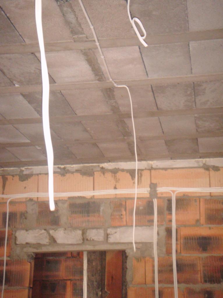 Instalacja elektryczna w surowym stanie: białe kable prowadzone po bloczkach i suficie, widoczne łączenia i fragment otworu okiennego. Prace w trakcie.