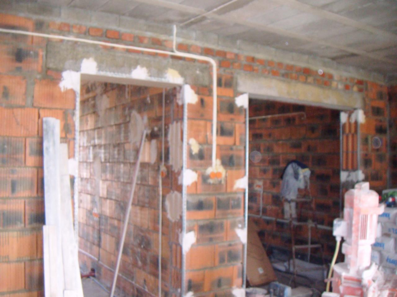 Surowy mur z cegły z widoczną instalacją elektryczną i przygotowanymi otworami na drzwi, w trakcie prac wykończeniowych, z widocznym sprzętem budowlanym.