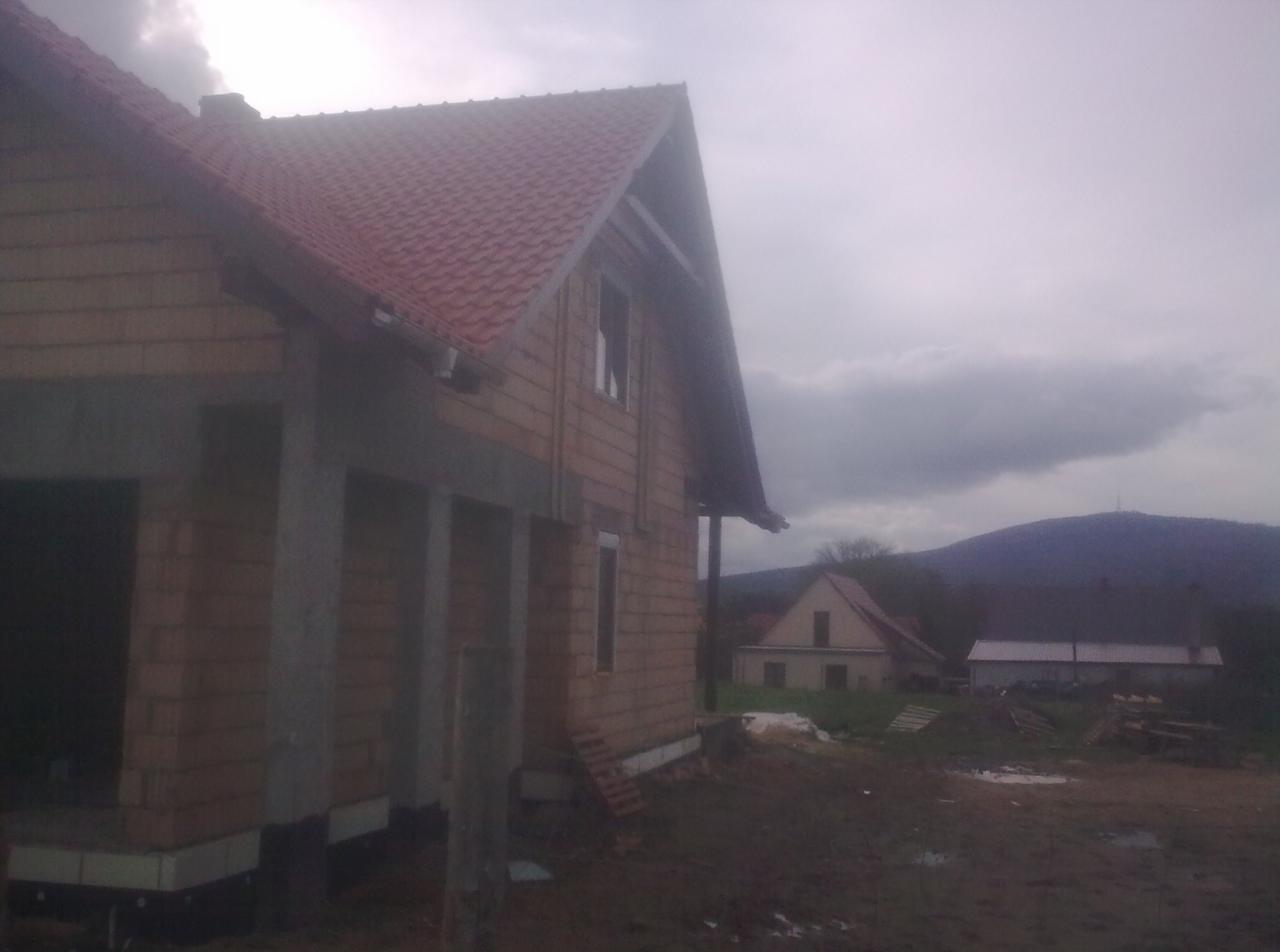 Surowy stan budynku z cegły z czerwonym dachem w pochmurny dzień. Widoczne elementy konstrukcyjne i zarys gór w tle. Etap budowy, widok z zewnątrz.