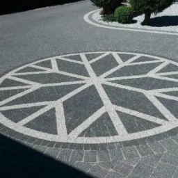 Szara kostka brukowa ułożona w okrągły wzór gwiazdy na podjeździe, z elementami dekoracyjnymi z białego kamienia i zadbaną zielenią w tle.
