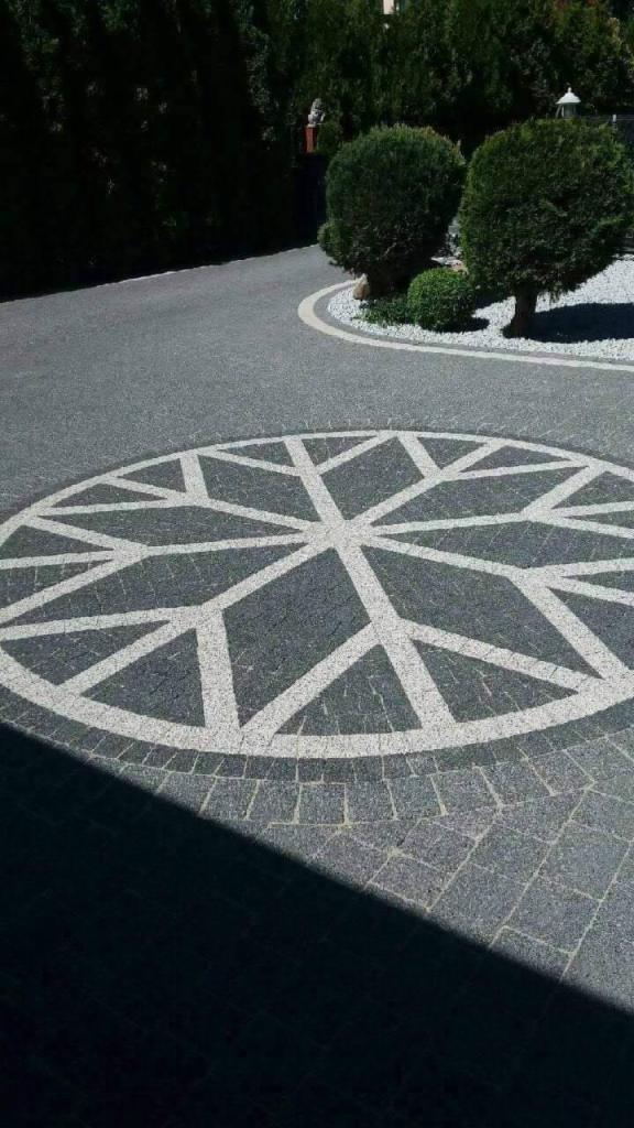 Szara kostka brukowa ułożona w okrągły wzór gwiazdy na podjeździe, z elementami dekoracyjnymi z białego kamienia i zadbaną zielenią w tle.