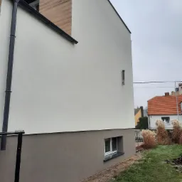 Fragment bocznej ściany budynku po nałożeniu dwukolorowej elewacji: górna część biała, dolna szara, z widoczną rynną i oknem piwnicznym, na tle trawnika i zabudowań sąsiednich.