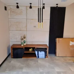 Wykończony salon z marmurową ścianą, drewnianą szafką RTV, czarnym panelem akustycznym i lampami wiszącymi w stylu industrialnym, karton po telewizorze oparty o ścianę.