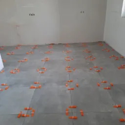 Ułożone szare płytki podłogowe z pomarańczowymi klipsami poziomującymi w trakcie prac wykończeniowych w jasnym pomieszczeniu.