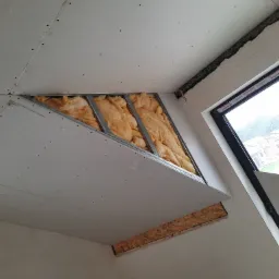 Montaż płyty gipsowo-kartonowej na suficie pod skosem, widoczna wełna mineralna w konstrukcji, częściowo zakryta płytą, drabina oparta o ścianę, okno z widokiem na zewnątrz.