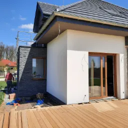 Nowoczesny dom jednorodzinny z fragmentem elewacji wykończonej białym tynkiem i szarym kamieniem dekoracyjnym, widoczne okna z drewnianą stolarką, częściowo wykonany drewniany taras i przygotowania...