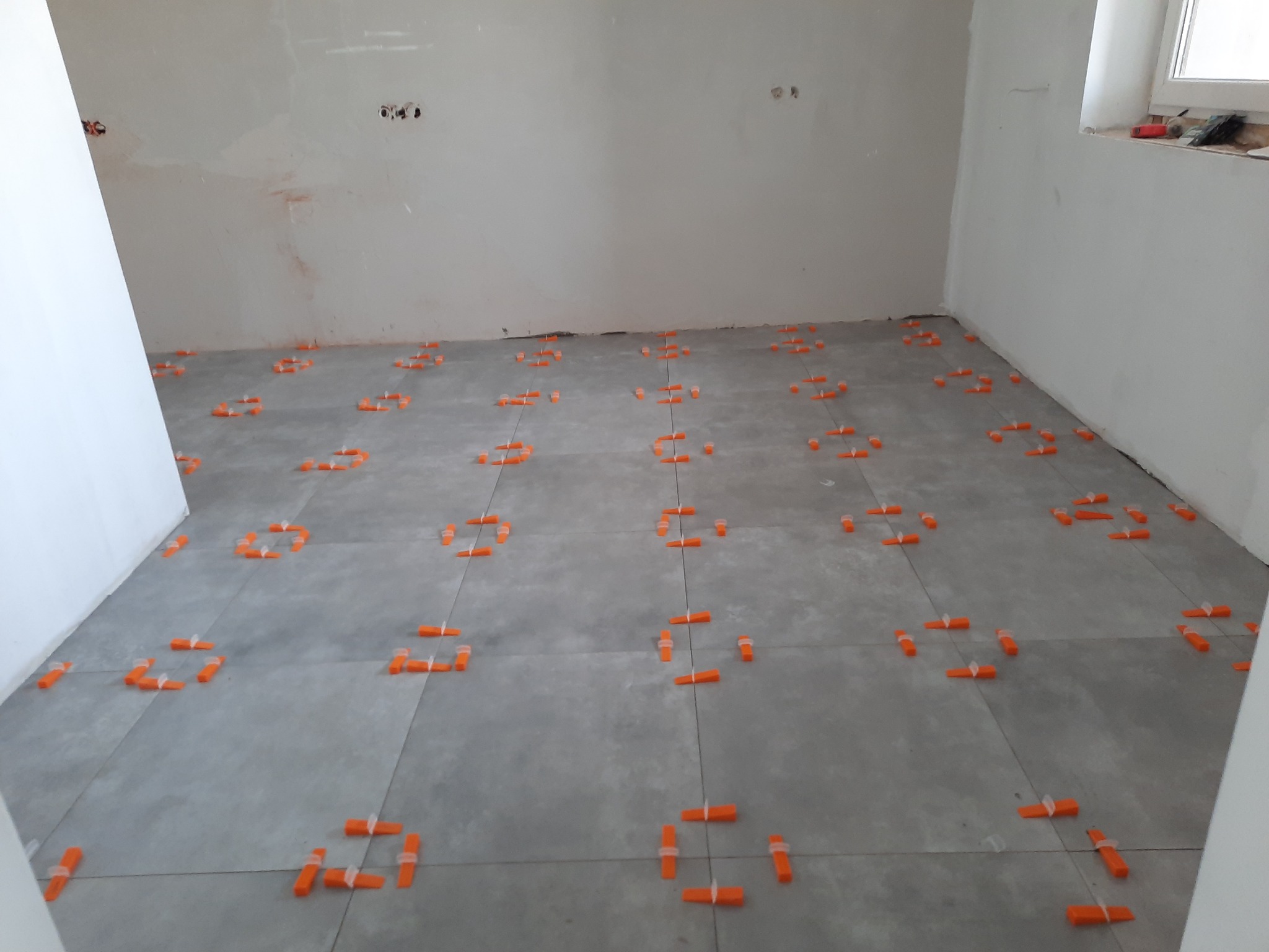 Ułożone szare płytki podłogowe z pomarańczowymi klipsami poziomującymi w trakcie prac wykończeniowych w jasnym pomieszczeniu.