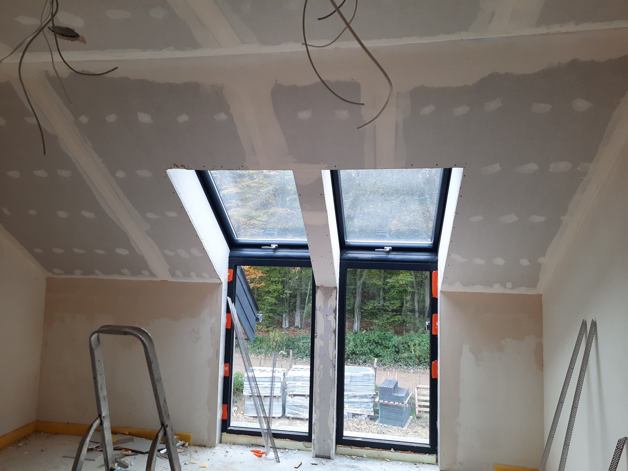 Poddasze w trakcie wykańczania: widoczne płyty karton gips na skośnym suficie, szpachlowane łączenia, okna dachowe z widokiem na jesienny las, drabina i materiały budowlane w pomieszczeniu.