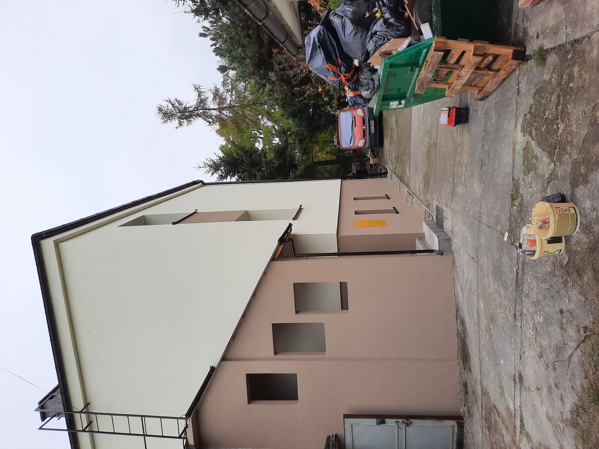 Dwukondygnacyjny budynek z częściowo odnowioną elewacją w odcieniach beżu i kremu, widoczny z boku, z ustawioną drabiną oraz wiadrami z farbą na chodniku, w tle kontener na śmieci i zaparkowany...