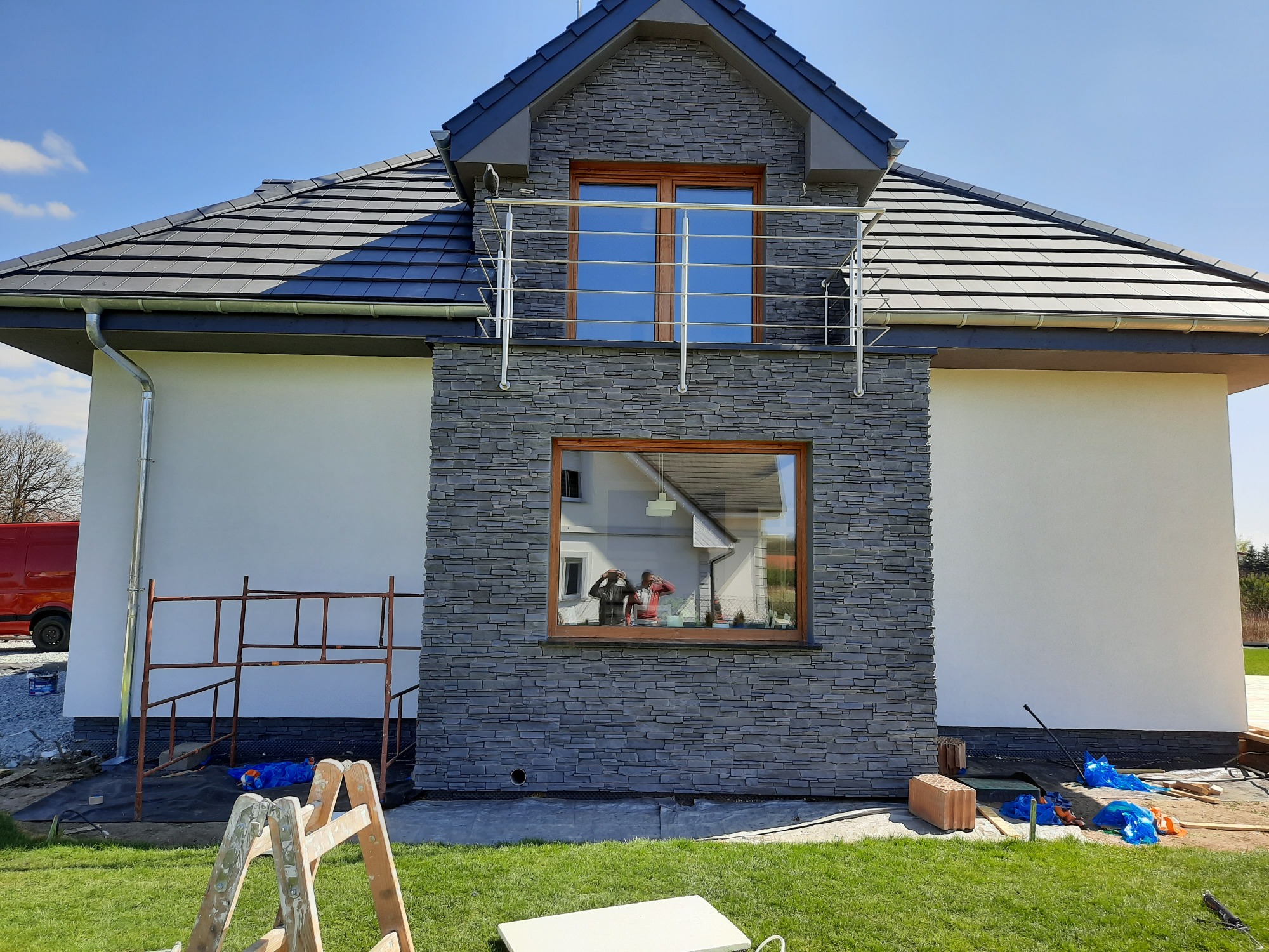 Nowoczesny dom jednorodzinny z fragmentem elewacji wykończonym szarym kamieniem dekoracyjnym, widoczne rusztowanie i narzędzia sugerujące prace wykończeniowe, odbicie dwóch osób w oknie.
