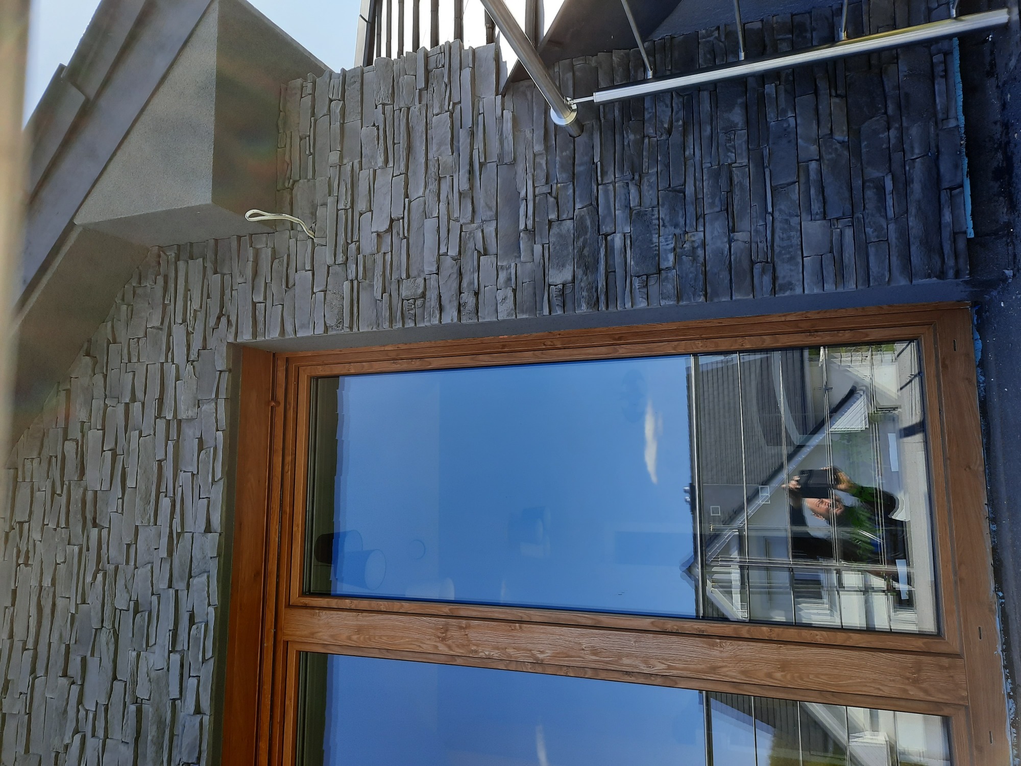 Fragment elewacji budynku wykończonej szarym kamieniem dekoracyjnym, widoczne drewniane obramowanie okna z odbiciem fotografa i otoczenia.