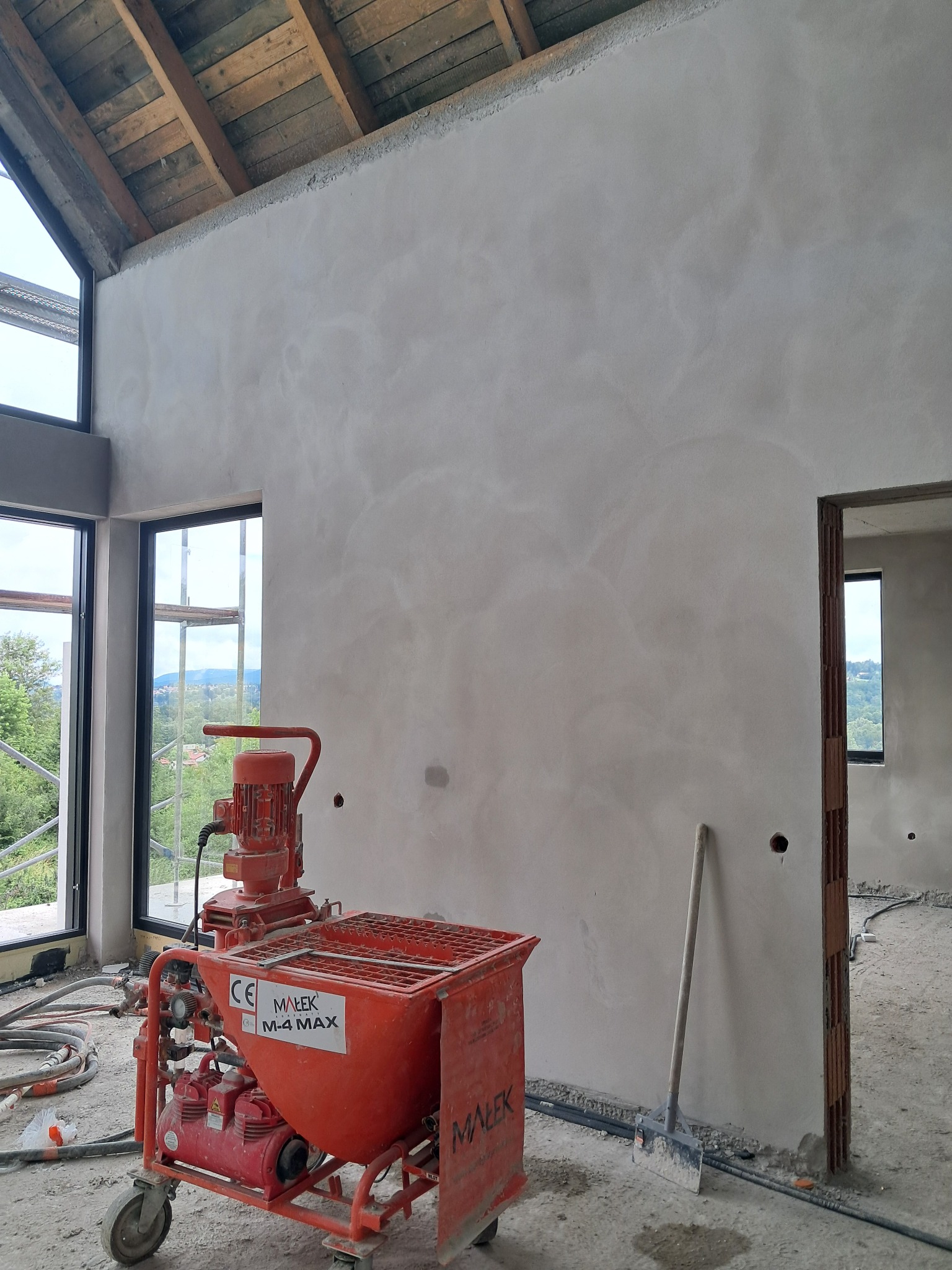 Świeżo otynkowana ściana w surowym wnętrzu z widoczną maszyną do tynkowania Malek M-4 MAX i szpachlą, duże okna z widokiem na zieleń.