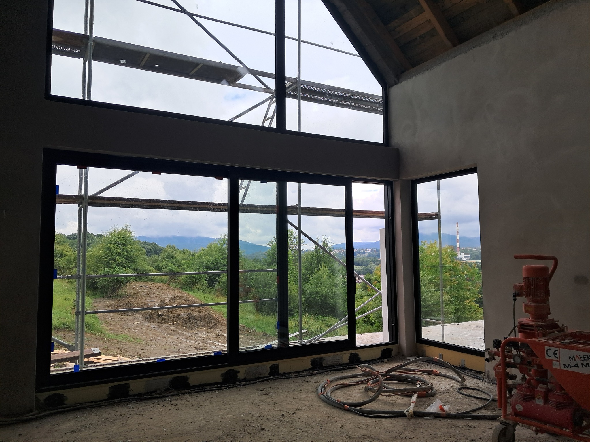 Wnętrze w trakcie tynkowania maszynowego z widokiem na zewnątrz przez duże okna. Widoczne rusztowania i maszyna do tynkowania.