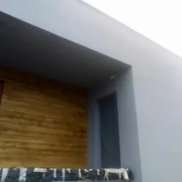 Fragment elewacji budynku z tynkiem strukturalnym imitującym drewno, zabezpieczona folią przed oknem, obok szara, gładka ściana.