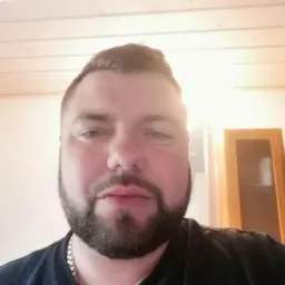 Portret brodatego mężczyzny w czarnej koszulce na tle jasnego sufitu z drewnianą boazerią i fragmentu szafy. Widoczne światło słoneczne.