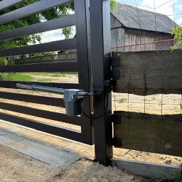 FHUP ORBI GRZEGORZ WISIŃSKI - Nowoczesna, brązowa brama wjazdowa z automatycznym napędem ramieniowym, widoczna na tle wiejskiej zabudowy z drewnianą stodołą.