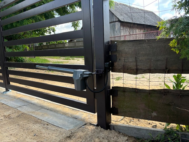 Nowoczesna, brązowa brama wjazdowa z automatycznym napędem ramieniowym, widoczna na tle wiejskiej zabudowy z drewnianą stodołą.