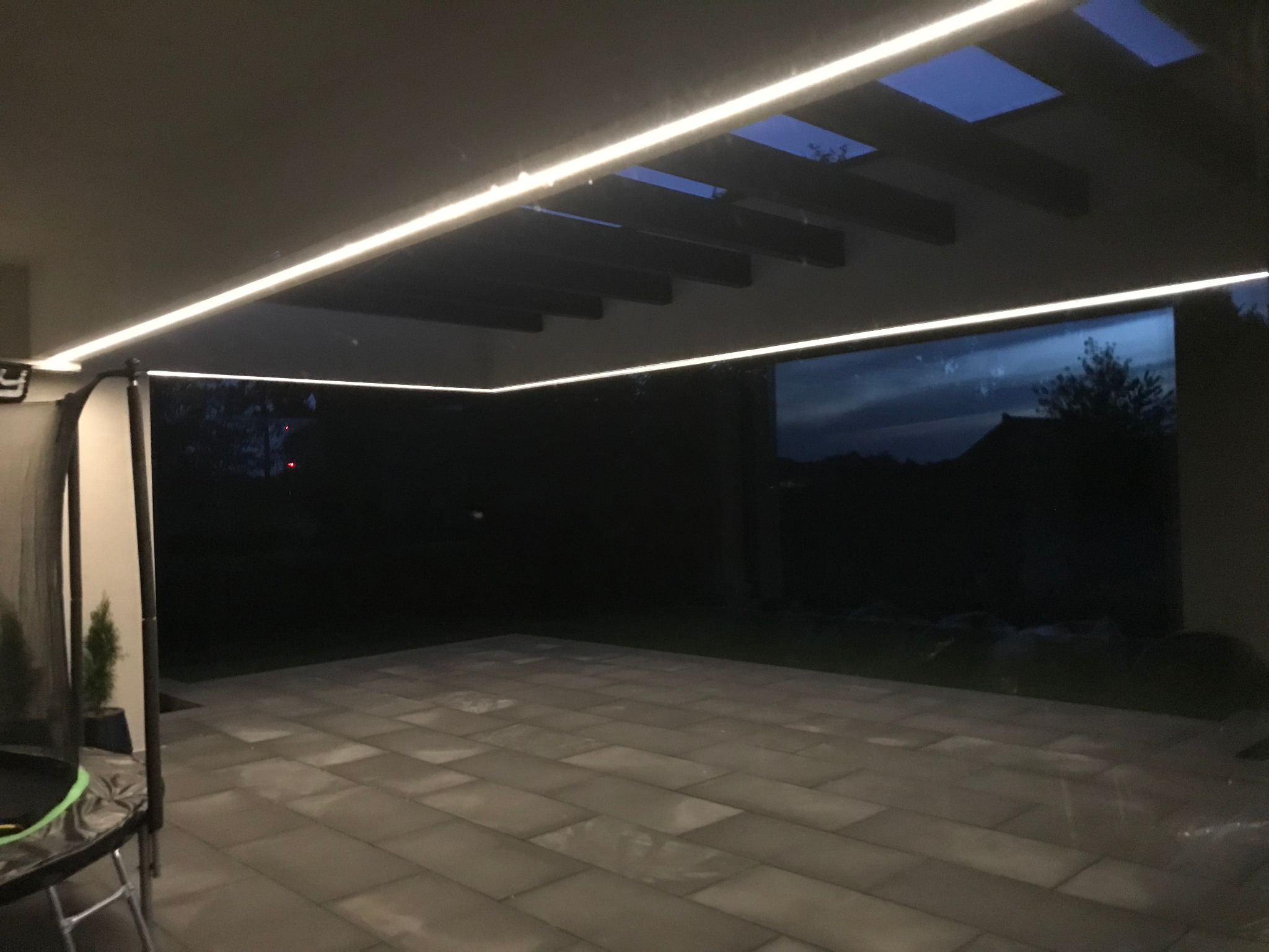 Nowoczesny taras z podświetlanym sufitem z drewnianych belek i liniowym oświetleniem LED, widok na nocny ogród.