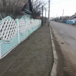Długi, dekoracyjny, betonowy płot pomalowany na biało i turkusowo, oddzielający posesję od ulicy z nowo wysypanym żwirem na chodniku.