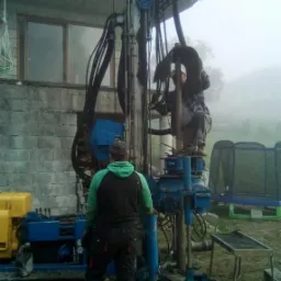 Dwóch mężczyzn obsługuje niebieską wiertnicę na błotnistym terenie, z budynkiem w tle w porannej mgle.