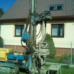 Wiertnica SATVIA VB 150R w trakcie pracy na tle domu jednorodzinnego z brązowym dachem i białym ogrodzeniem. Widoczny osprzęt wiertniczy i fragment wykopu.