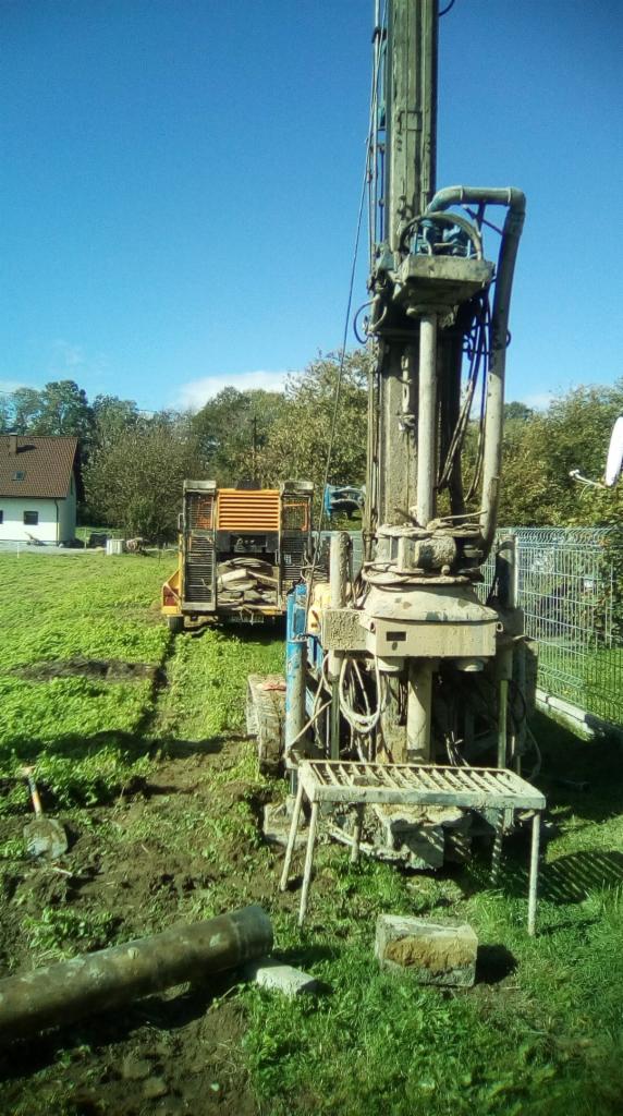 Wiertnica w trakcie pracy na trawiastym terenie, widoczny sprzęt pomocniczy i fragment rury, w tle dom jednorodzinny.