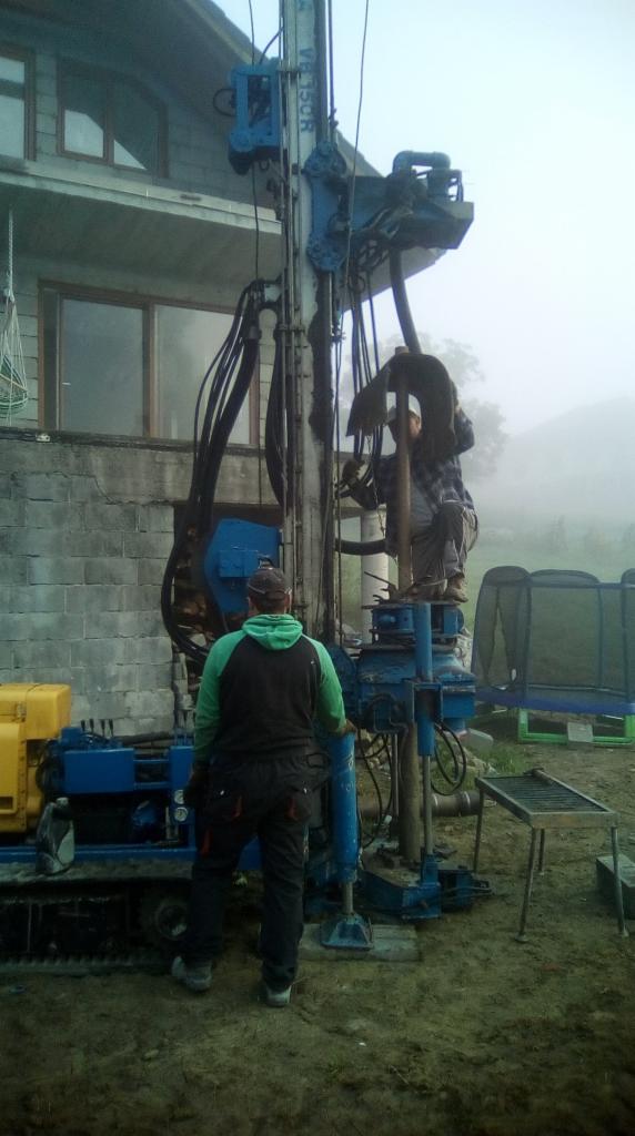 Dwóch mężczyzn obsługuje niebieską wiertnicę na błotnistym terenie, z budynkiem w tle w porannej mgle.