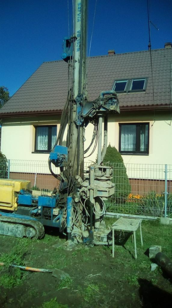 Wiertnica SATVIA VB 150R w trakcie pracy na tle domu jednorodzinnego z brązowym dachem i białym ogrodzeniem. Widoczny osprzęt wiertniczy i fragment wykopu.