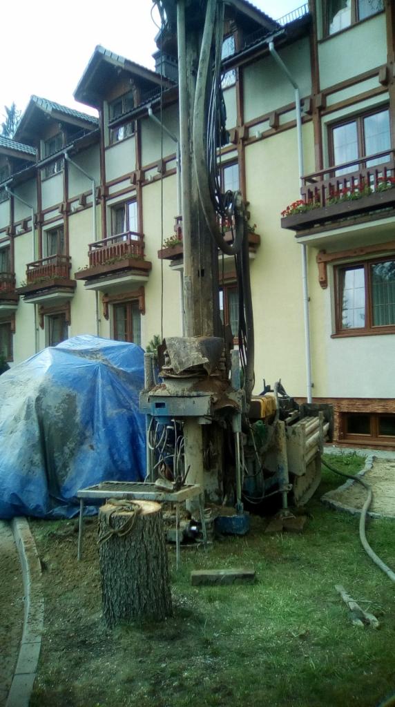 Wiertnica do studni głębinowej na tle budynku z balkonami z kwiatami, przykryta plandeką część sprzętu, obok pniak z narzędziami.