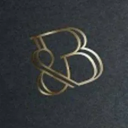 Złoty, stylizowany monogram 'B&' na ciemnym, matowym tle, oświetlony z góry.