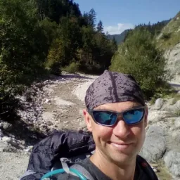 Uśmiechnięty mężczyzna w okularach przeciwsłonecznych i bandanie na głowie, z plecakiem turystycznym na tle górskiego potoku i lasu.