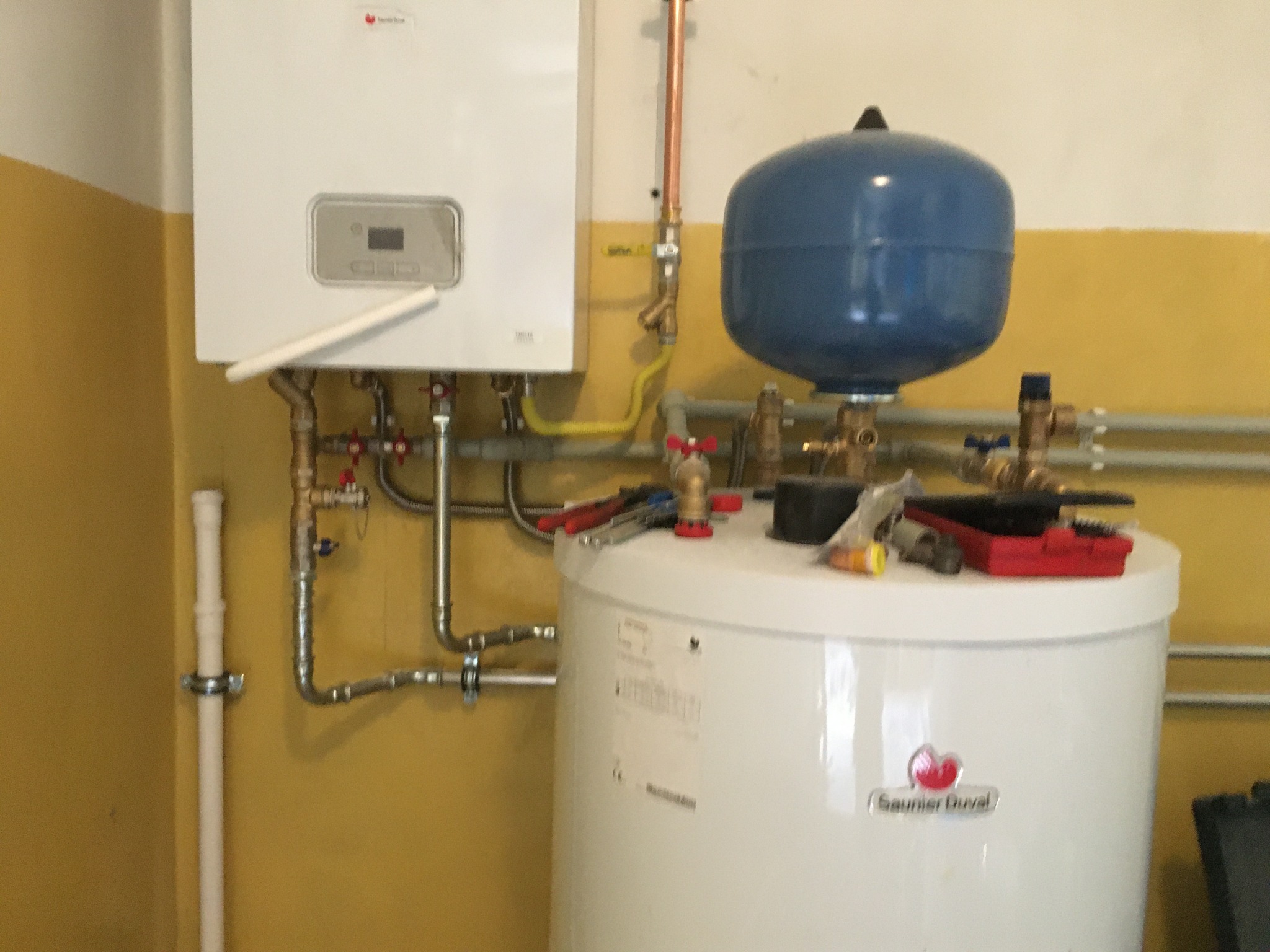 Biały piec gazowy Saunier Duval z niebieskim zbiornikiem wyrównawczym, widoczne rury miedziane i stalowe, narzędzia na górze zbiornika, ściana pomalowana na żółto-biało.