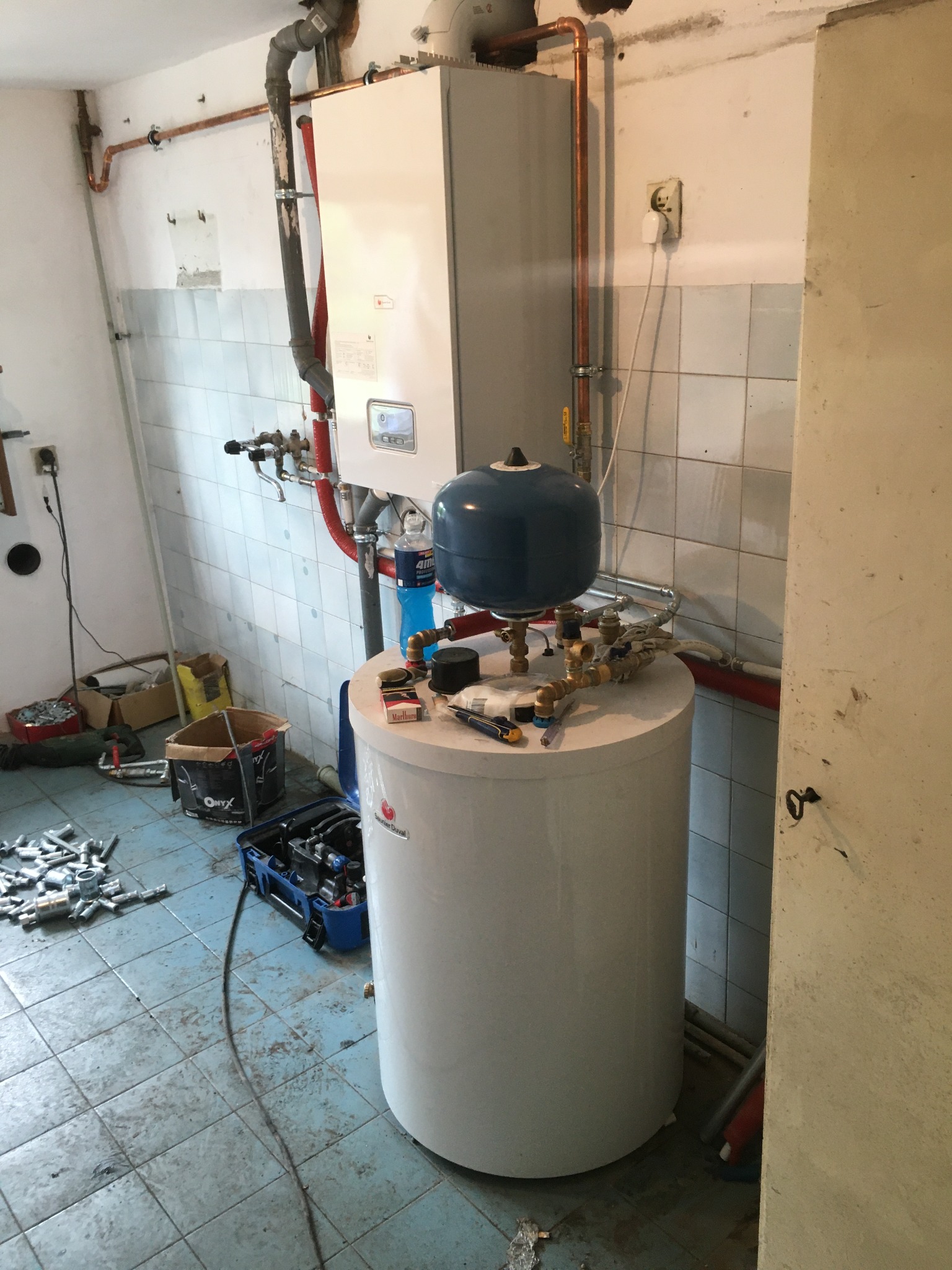 Bojler z niebieską głowicą podłączony do instalacji CO, narzędzia i materiały robocze na wierzchu, widoczne rury miedziane, płytki ceramiczne na ścianach, pomieszczenie w trakcie prac instalacyjnych.