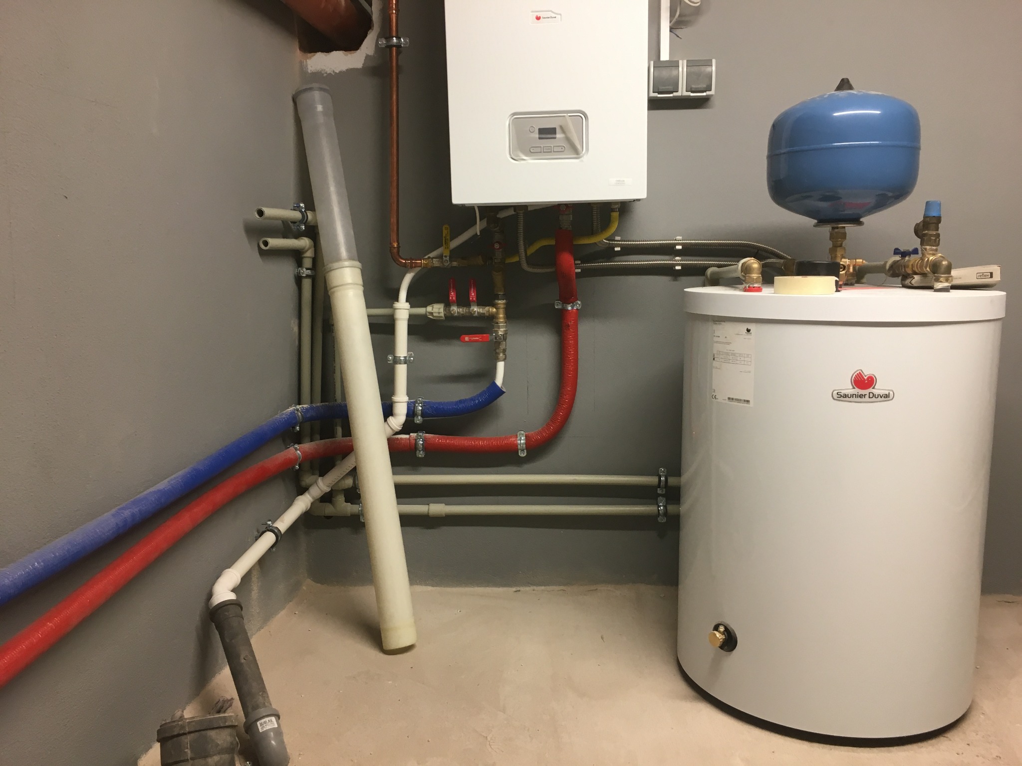 Biały piec gazowy Saunier Duval z podłączonymi rurami miedzianymi i plastikowymi w kolorach czerwonym i niebieskim, zbiornik wyrównawczy z niebieską górną częścią, szare ściany i podłoga betonowa.