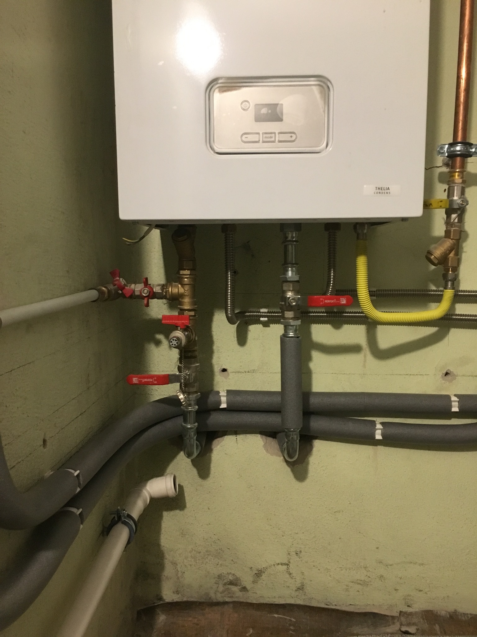 Biały piec gazowy Thelia Condens zamontowany na ścianie z widocznymi rurami gazowymi, zaworami z czerwonymi rączkami oraz izolowanymi rurami grzewczymi.