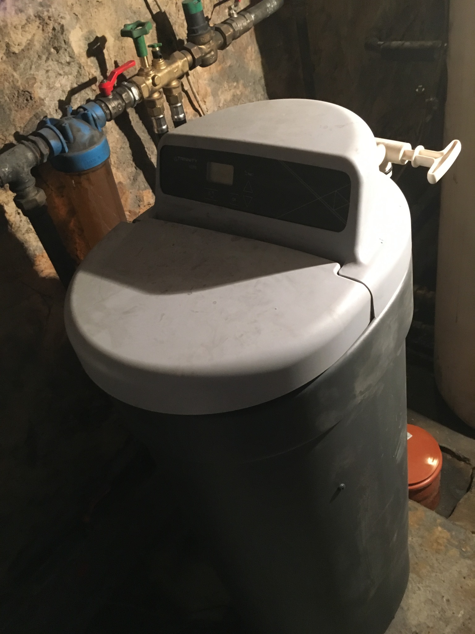 Szara centralna jednostka uzdatniania wody z widocznymi rurami i zaworami na tle surowej ściany piwnicy.