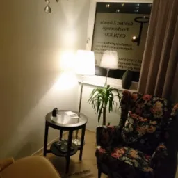 Wnętrze gabinetu w Sosnowcu z kryształowym żyrandolem, dwoma fotelami (jeden w kwiatowy wzór, drugi żółty), stolikiem z lampą i rośliną w doniczce przy oknie z widocznym napisem 'exbrico'.