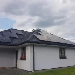 Dom jednorodzinny z panelami słonecznymi na czarnym, metalowym dachu, widok z poziomu trawnika w pochmurny dzień.