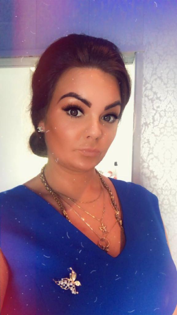 Portret kobiety w niebieskiej sukience z broszką motyla i złotą biżuterią na tle wzorzystej ściany. Widoczny fragment lustra.