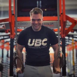 Uśmiechnięty mężczyzna w czarnej koszulce UBC trzyma się kółek gimnastycznych na tle pomarańczowej konstrukcji w siłowni.